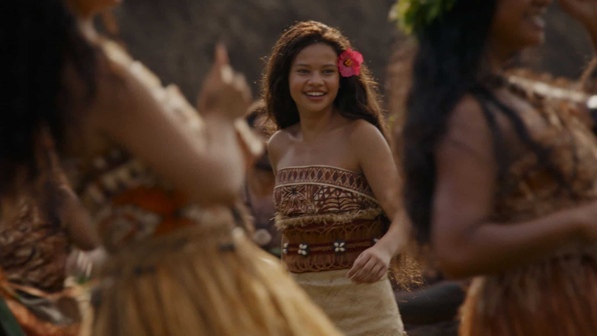 A young woman with long hair and a pink flower behind her ear smiles while dancing outdoors with others in traditional attire.
