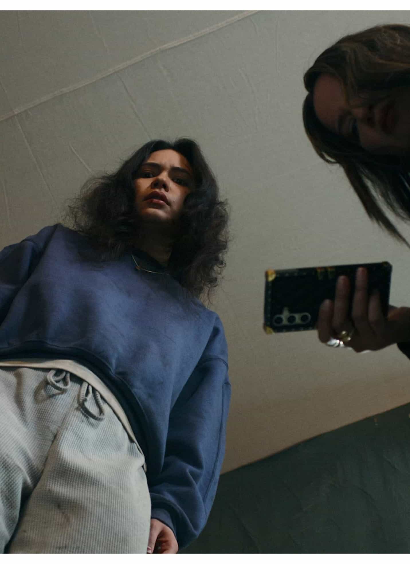 Two people look down towards the camera, one holding a phone and the other with a serious expression, both appearing to peer into a box or container. The scene is shot from a low angle.