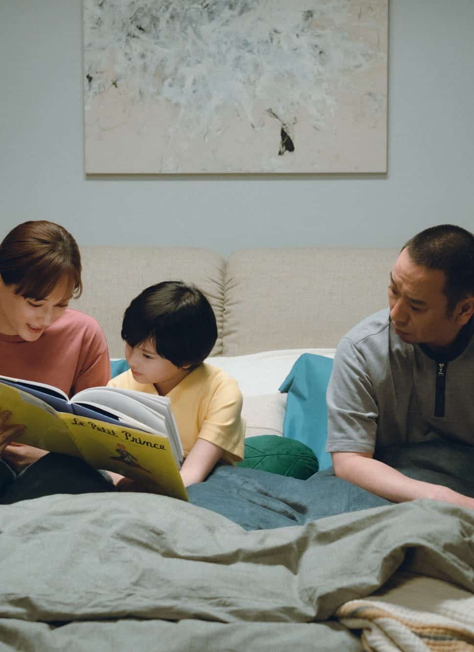 A woman reads a book to a child while a man sits beside them on a bed. They are under blankets in a softly lit bedroom with a painting on the wall and a lamp on a nightstand.