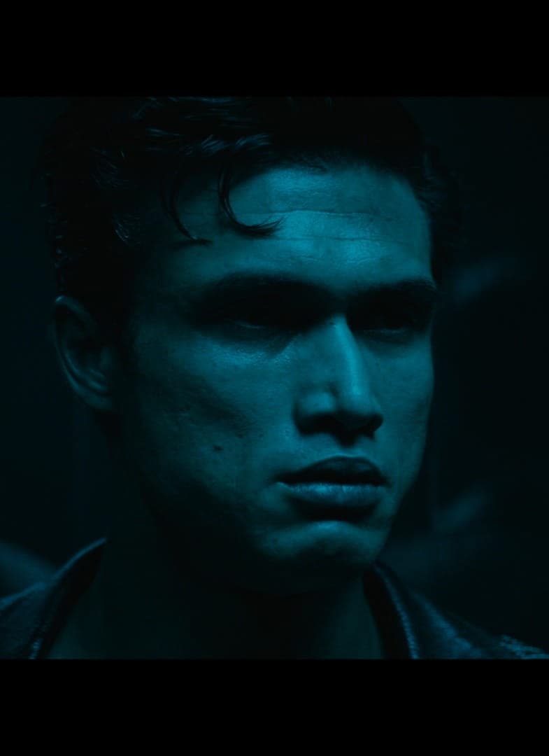 A young man with short dark hair stands in dim blue light, his expression serious. Shadows and reflections are visible behind him, creating a mysterious atmosphere.