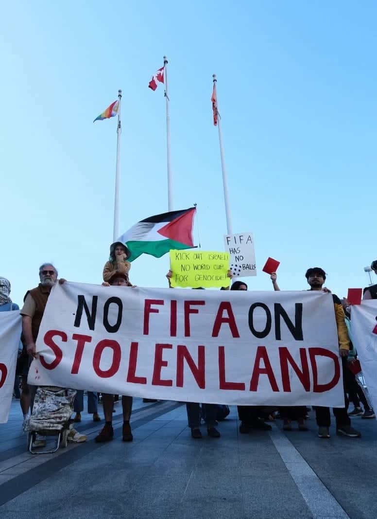 A group of protesters holds banners reading “NO FIFA ON STOLEN LAND” and “SHOW FIFA THE RED CARD,” with flags and tall buildings in the background.