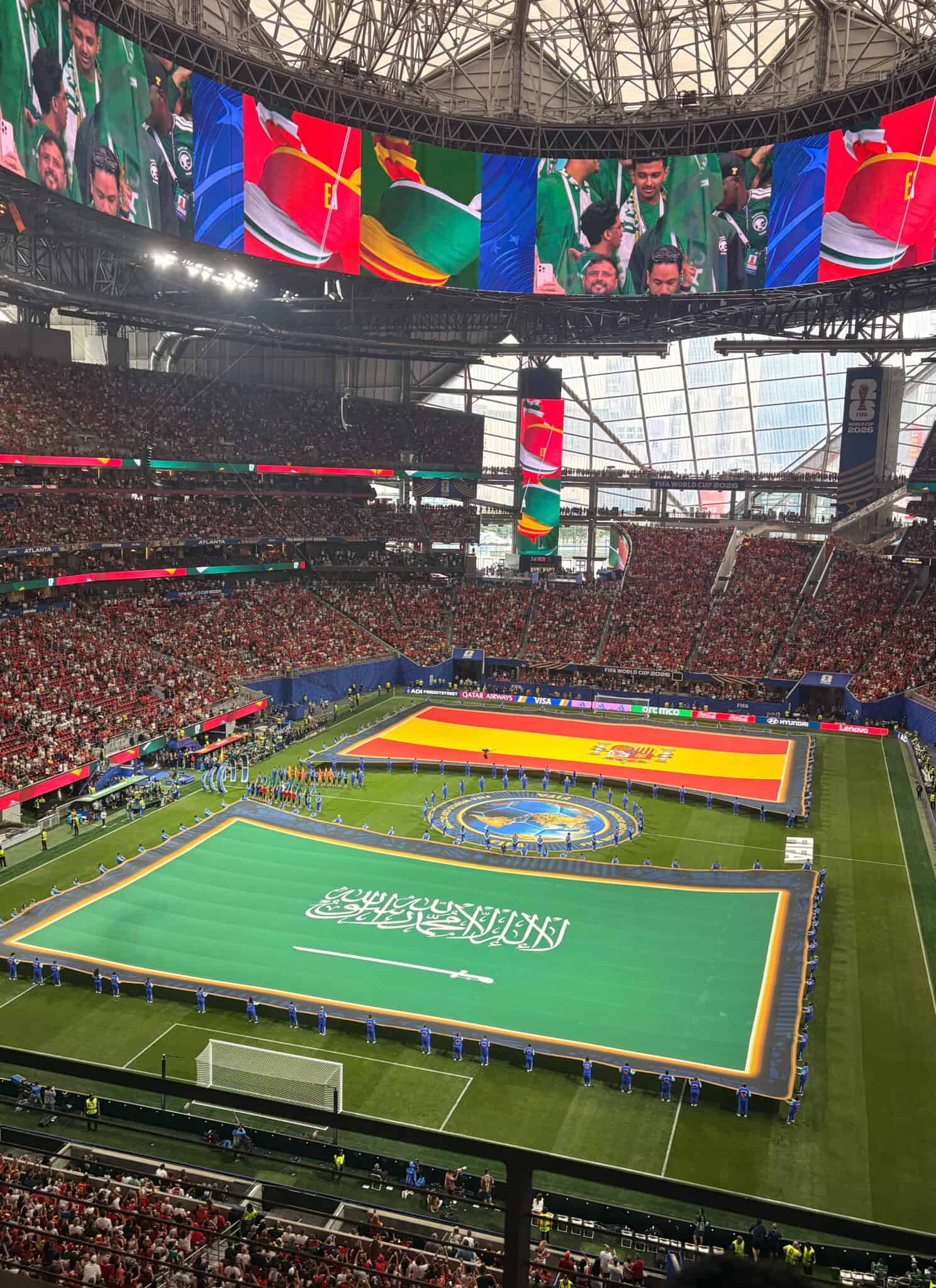Large flags of Saudi Arabia and Spain are displayed on a soccer field, surrounded by officials and a stadium full of spectators before a match.