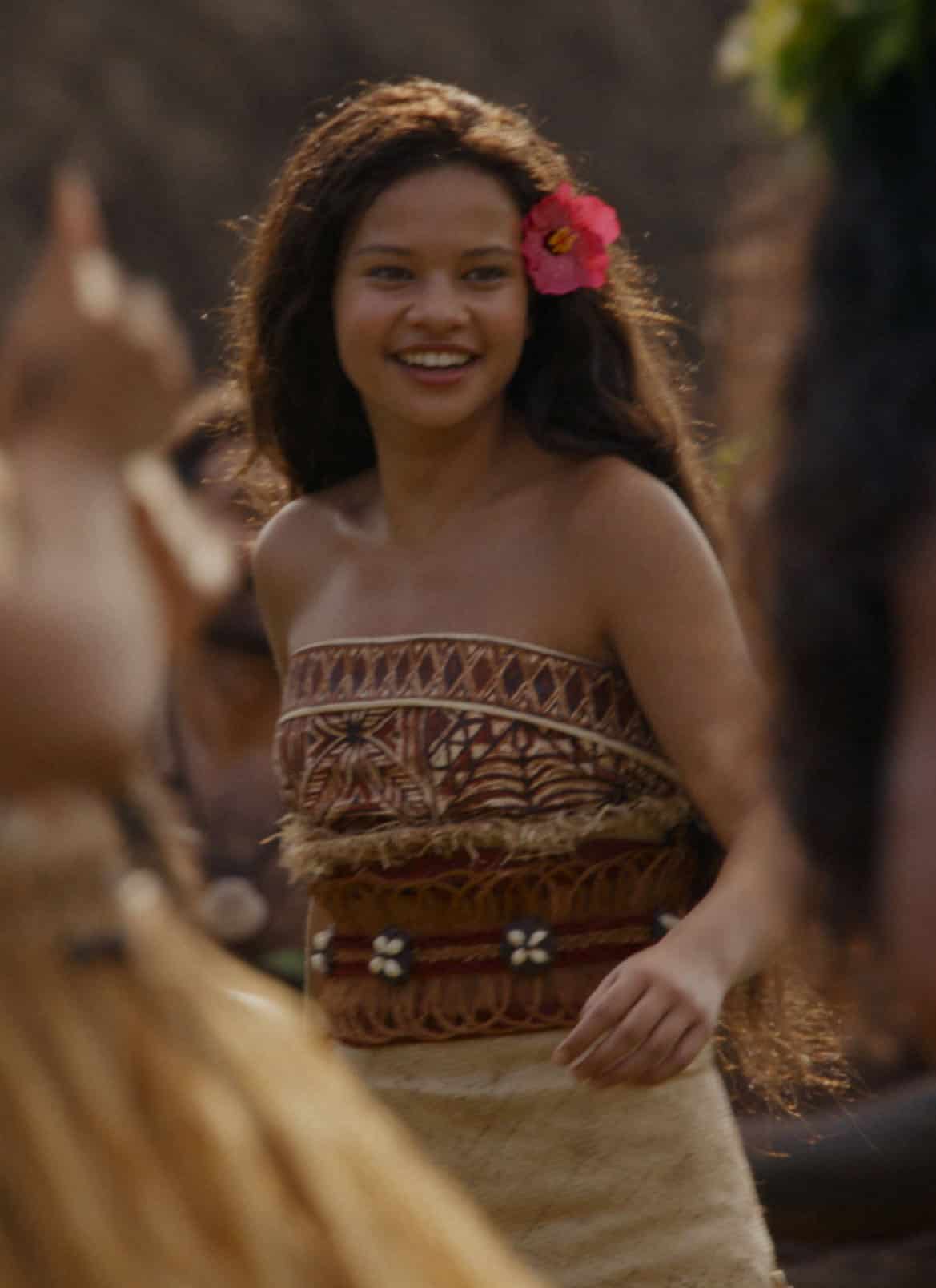 A young woman with long hair and a pink flower behind her ear smiles while dancing outdoors with others in traditional attire.