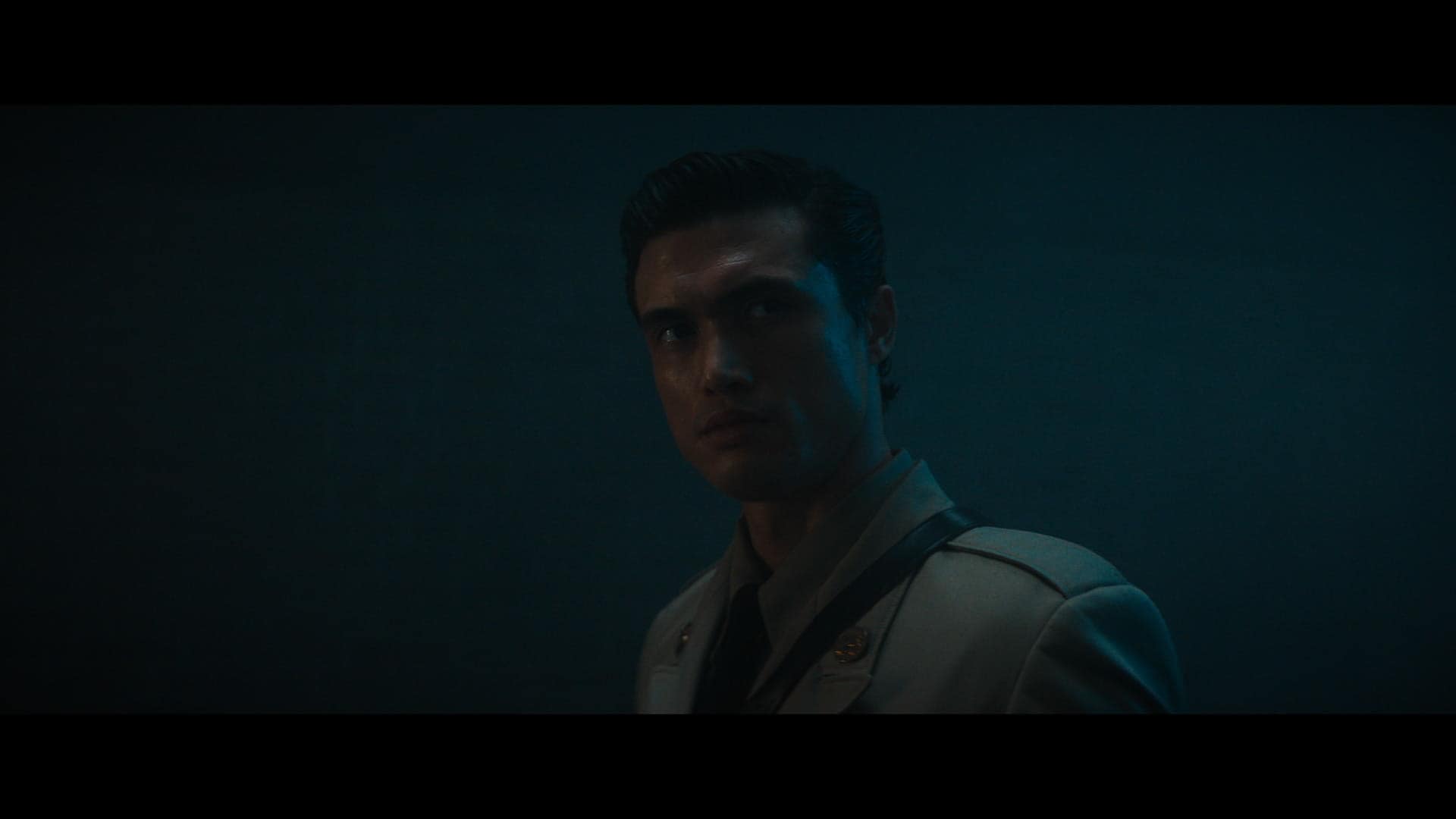 A man in a military-style uniform stands in dim, bluish lighting, looking off to the side with a serious expression.