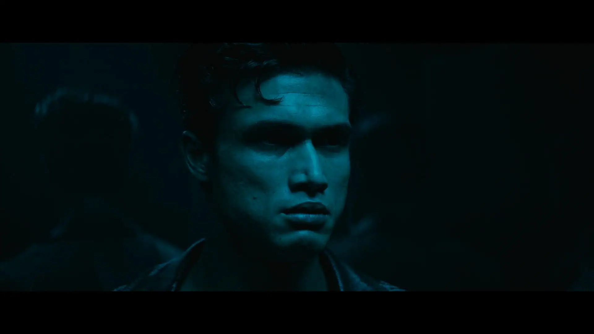 A young man with short dark hair stands in dim blue light, his expression serious. Shadows and reflections are visible behind him, creating a mysterious atmosphere.