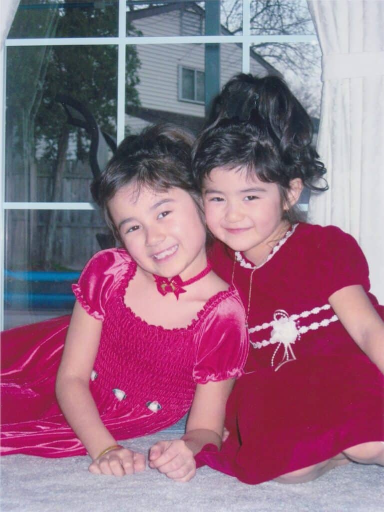 Two young Wasian girls in matching red velvet dresses sit and smile on a carpet in front of a large window, with houses and trees visible outside. Both have dark hair styled up and look happy and close together.