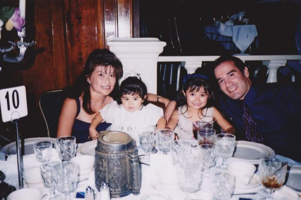A Wasian family of four sits at a round table set for a meal, smiling at the camera. The table is covered with clear glasses, plates, and a number 10 table sign. The background shows wood paneling and ornate decor.