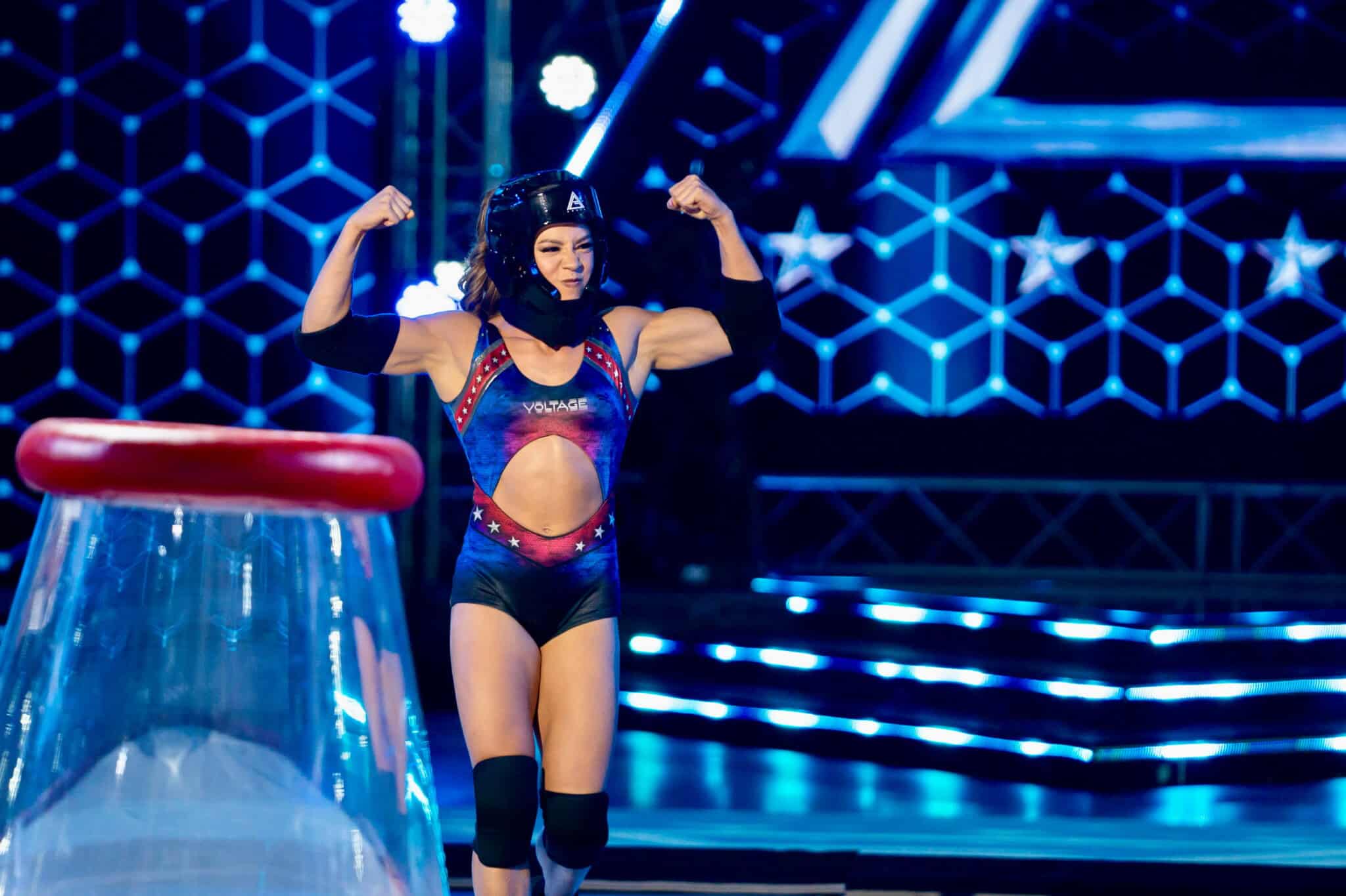 A female athlete in a helmet and sports gear flexes her arms confidently on a brightly lit competition stage with a futuristic background.
