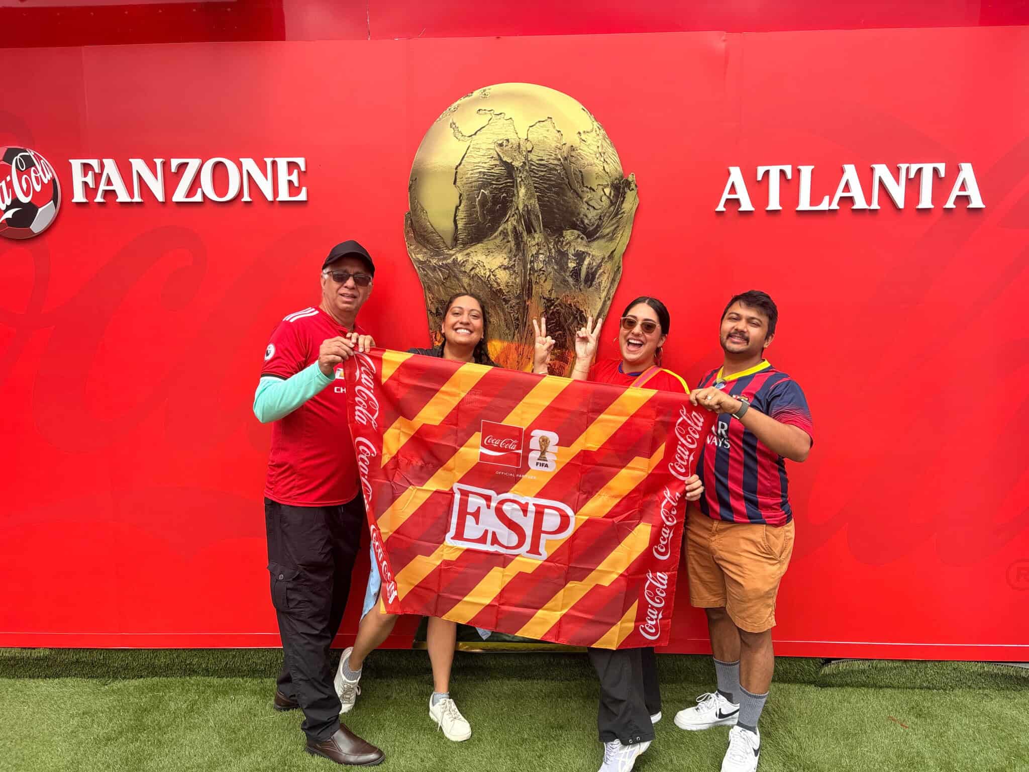 Four people smile and pose in front of a large World Cup trophy display. They hold a red and yellow ESP flag. The background reads FANZONE ATLANTA on a red wall with Coca-Cola branding.
