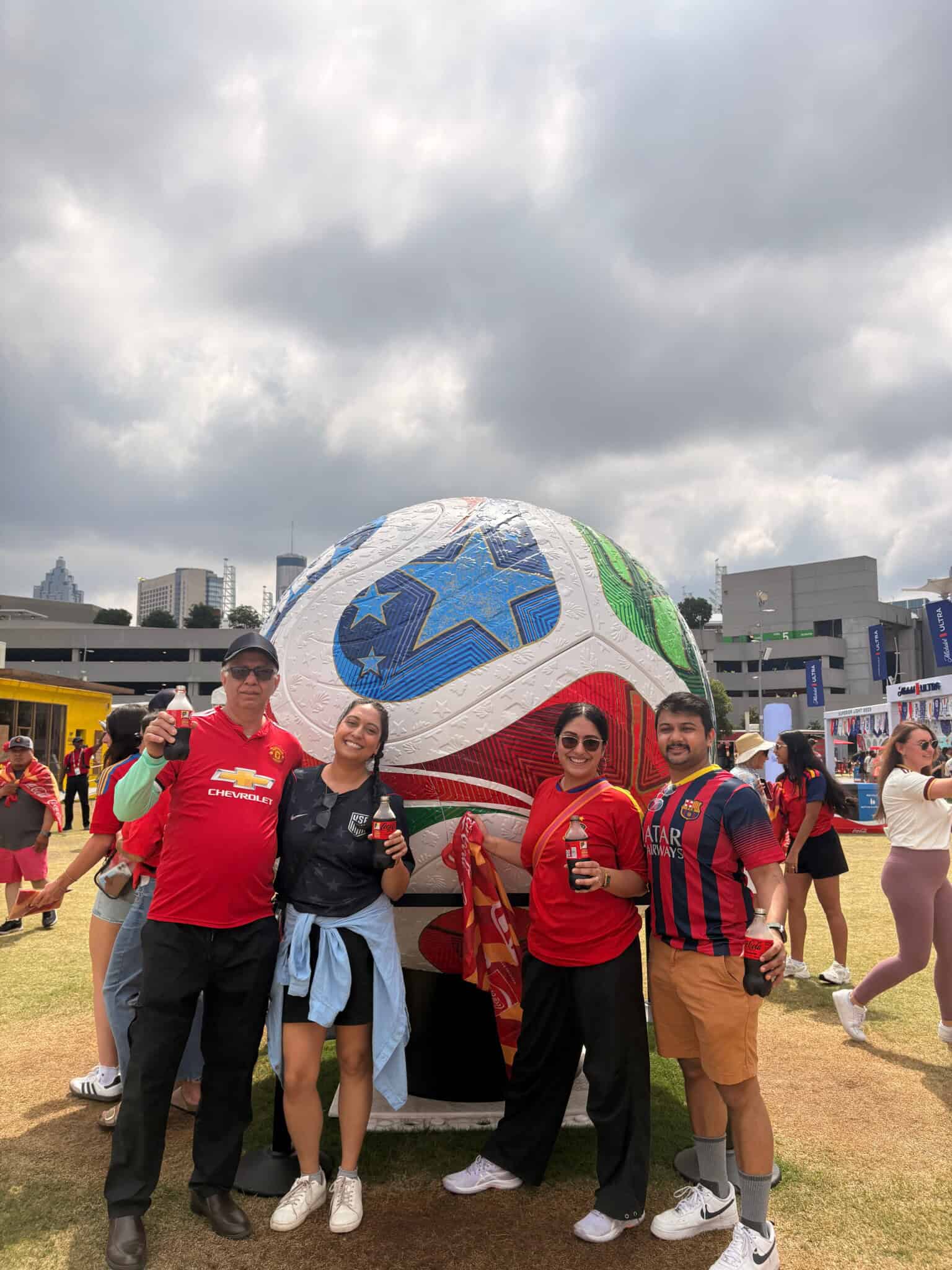 Four people pose smiling in front of a large soccer ball sculpture at an outdoor event, each holding drinks. They wear different soccer jerseys, with buildings and other attendees visible in the background under a cloudy sky.
