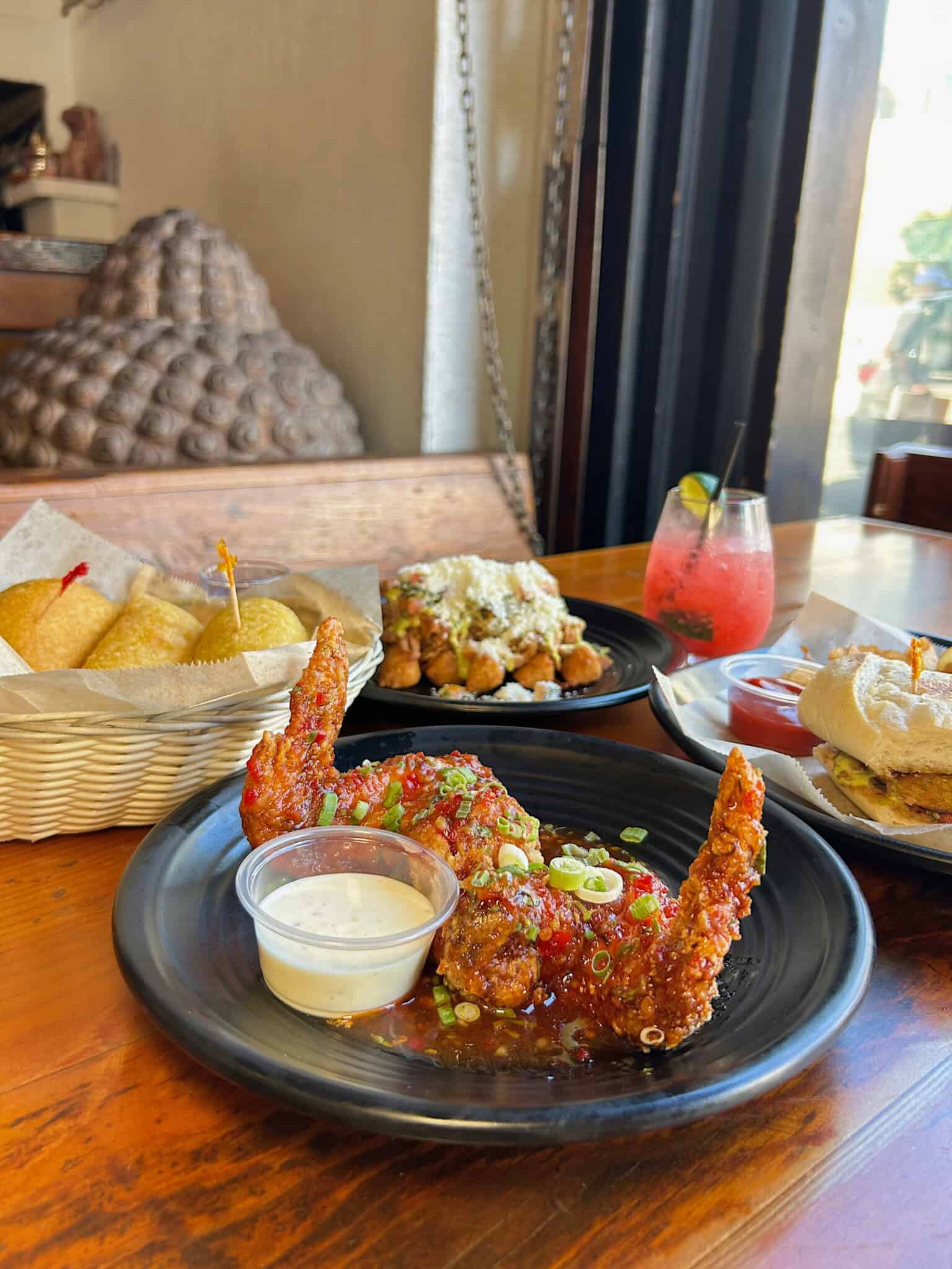 A plate of glazed fried chicken wings with dipping sauce, a basket of bread, a sandwich, loaded fries, and a red iced drink on a wooden table by a window.
