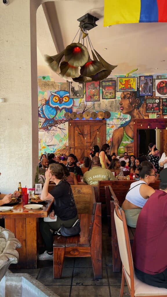 A lively restaurant scene with people dining at wooden tables, colorful wall art featuring an owl and a woman, and vibrant record covers decorating the background.