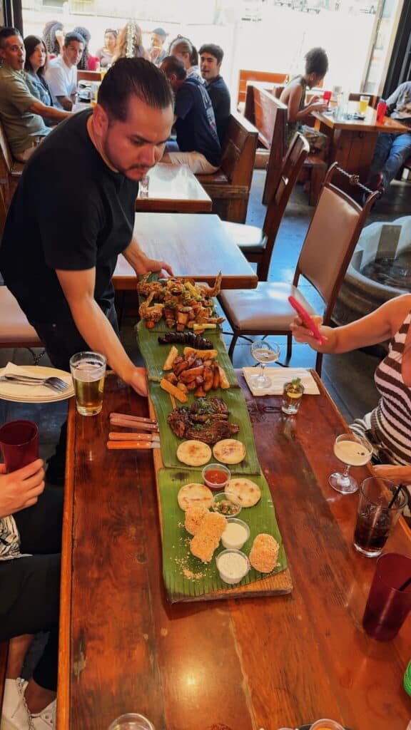 A server presents a long wooden platter covered with various foods on banana leaves to diners at a restaurant; people are seated around the table with drinks, ready to eat.