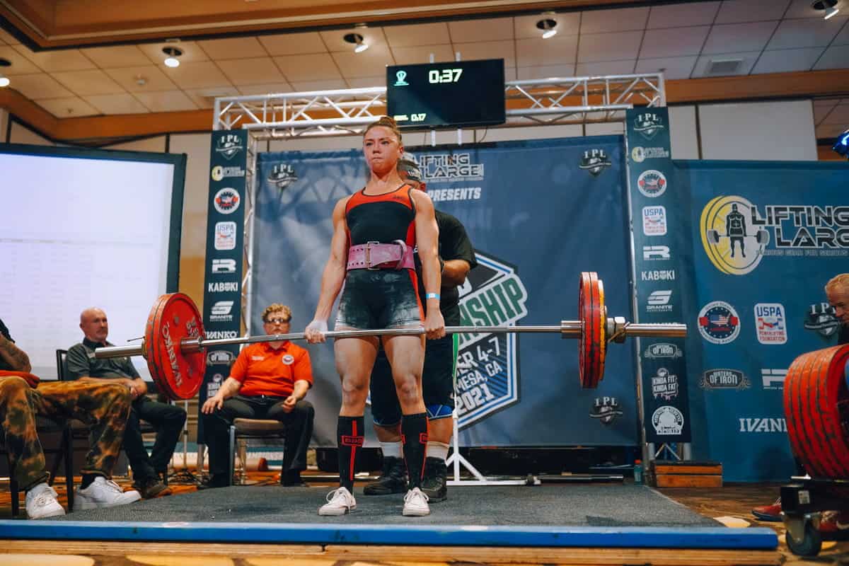 A female powerlifter in gym attire lifts a heavy barbell during a competition, with judges seated nearby and banners in the background.
