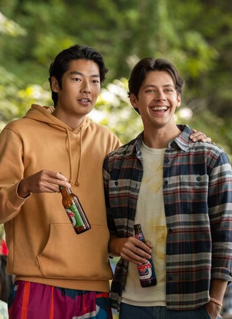 Two young men, including Joseph Chiu, stand close together at an outdoor gathering, both smiling and holding bottled drinks. One wears an orange hoodie, the other a plaid shirt. Other people mingle in the background amid greenery.