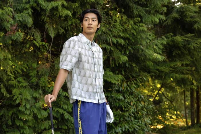 A man stands outdoors in front of leafy green trees, holding a golf club in his right hand. He wears a patterned short-sleeve shirt, blue athletic pants, and looks off to the side with a calm expression.