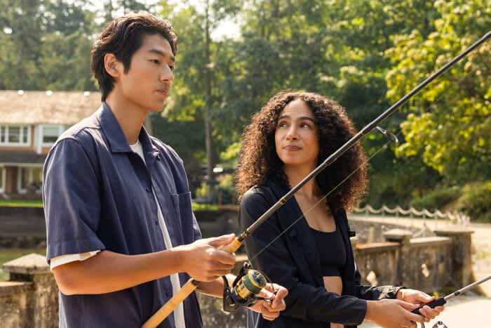 Two people stand outdoors holding fishing rods. The person on the left looks ahead while the person on the right, with curly hair, looks at them and smiles. Trees and a house are visible in the background.