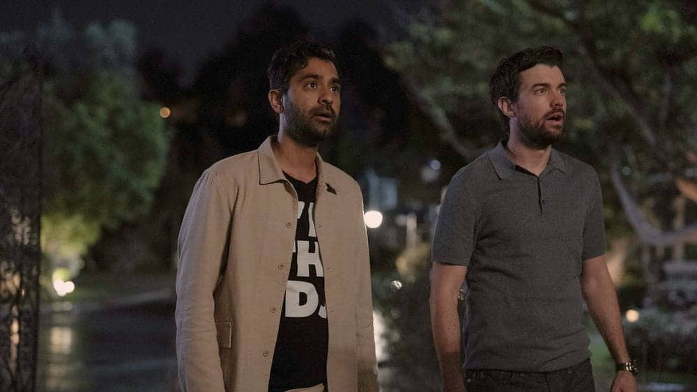 Two men stand outdoors at night, looking surprised or shocked. One wears a light jacket over a black shirt with white print, and the other wears a gray polo shirt. Trees and blurred lights are visible in the background.