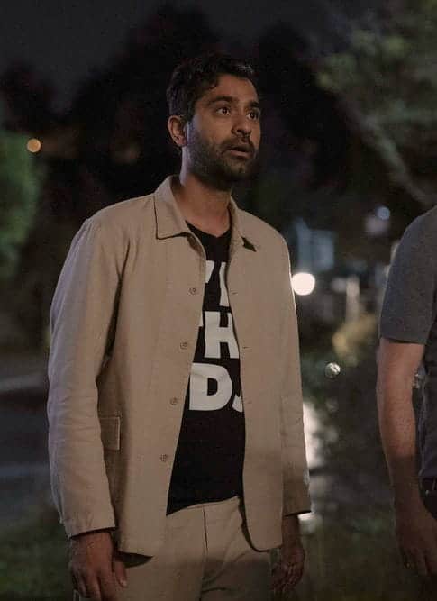 Two men stand outdoors at night, looking surprised or shocked. One wears a light jacket over a black shirt with white print, and the other wears a gray polo shirt. Trees and blurred lights are visible in the background.