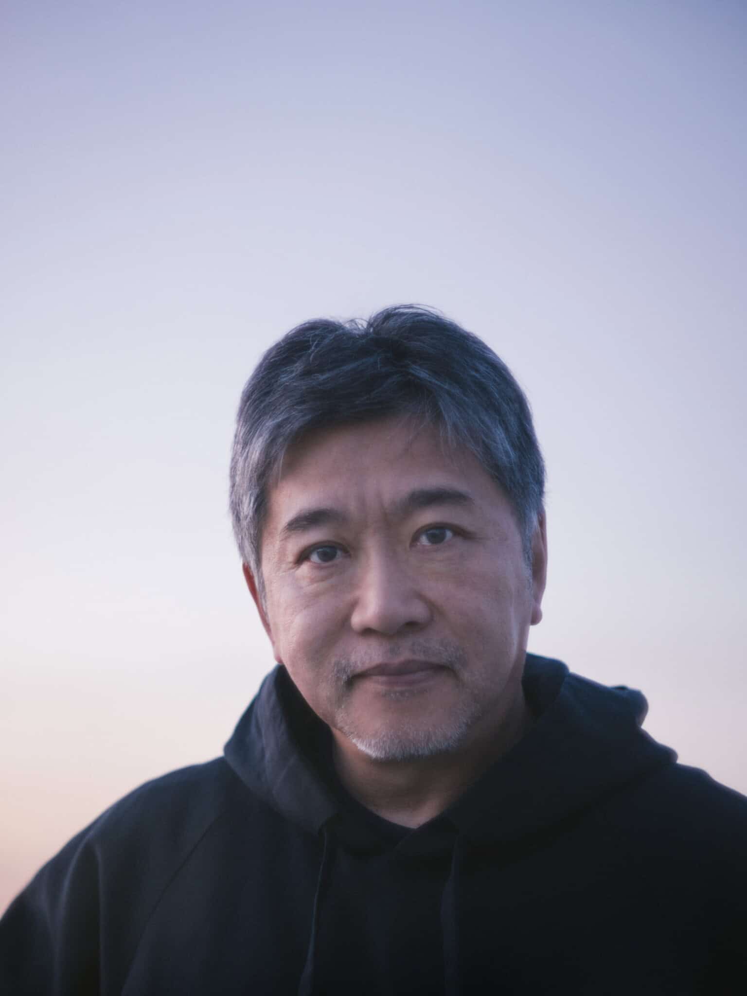 A middle-aged man with short gray hair and a trimmed beard wears a black hoodie, looking at the camera with a gentle expression. The background features a clear sky with soft, pastel colors at sunset.