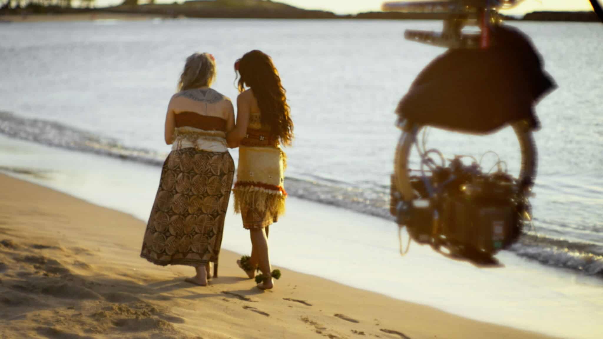 Two women in skirts walk along a sandy beach by the water, viewed from behind. A film camera is visible in the foreground, capturing the scene at sunset.