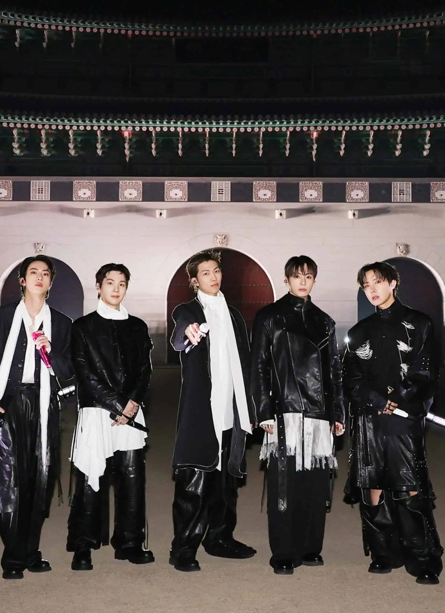 Seven men in stylish black outfits stand in a row at night in front of a large, traditional Korean building with multiple arches and an ornate tiled roof. The scene is well-lit with spotlights on the ground.