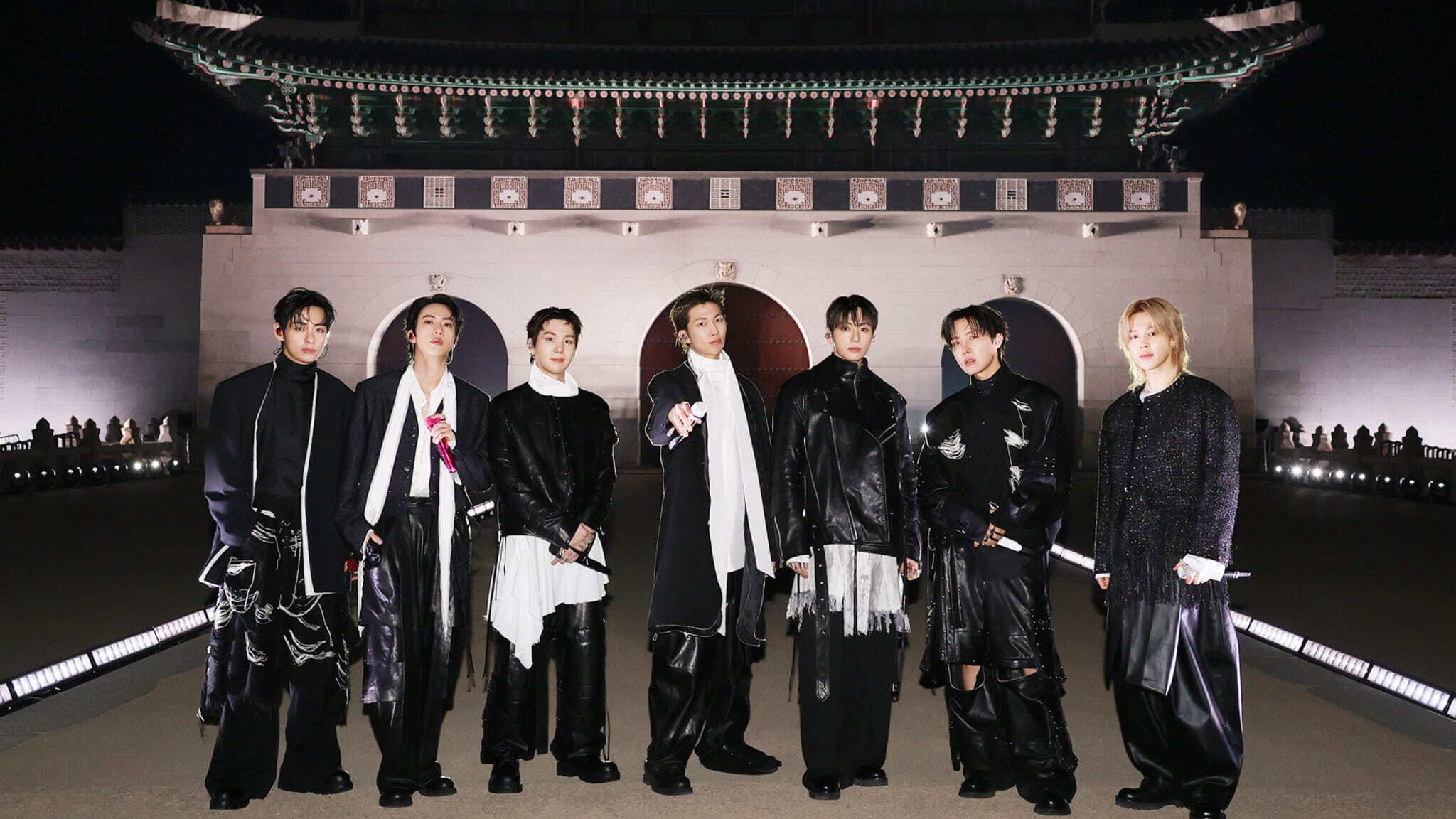 Seven men in stylish black outfits stand in a row at night in front of a large, traditional Korean building with multiple arches and an ornate tiled roof. The scene is well-lit with spotlights on the ground.
