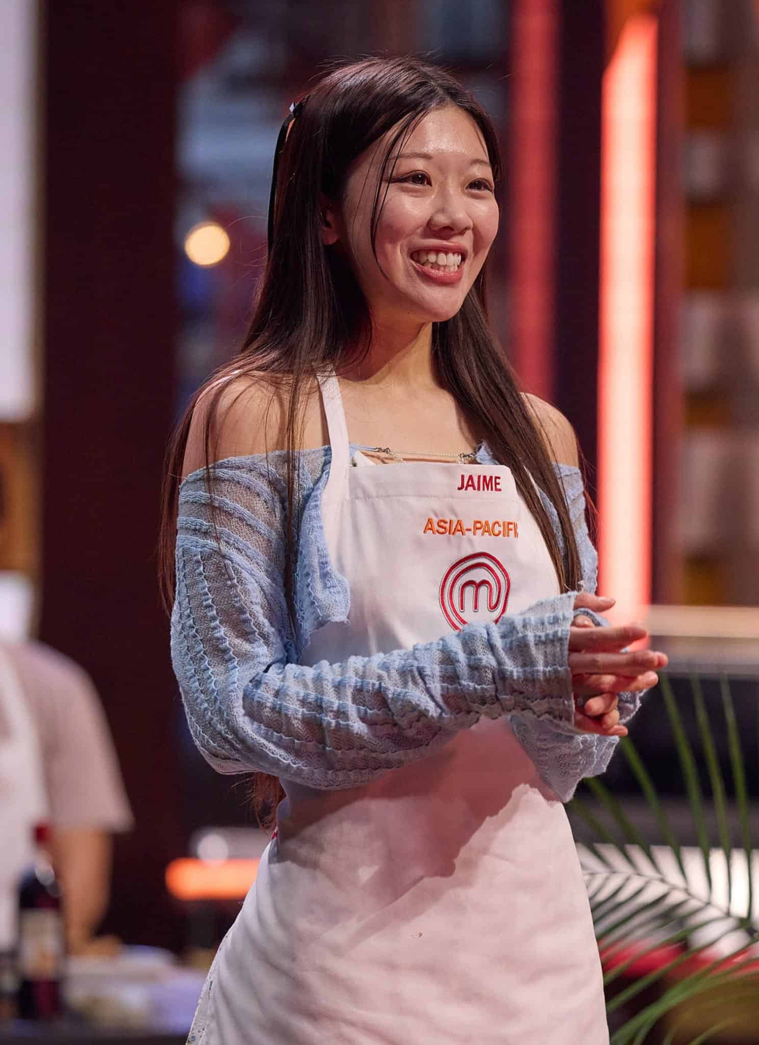 A smiling woman wearing a MasterChef Asia-Pacific apron stands indoors with hands clasped, radiating the confidence of a true Masterchef, while a blurred person sits in the background.