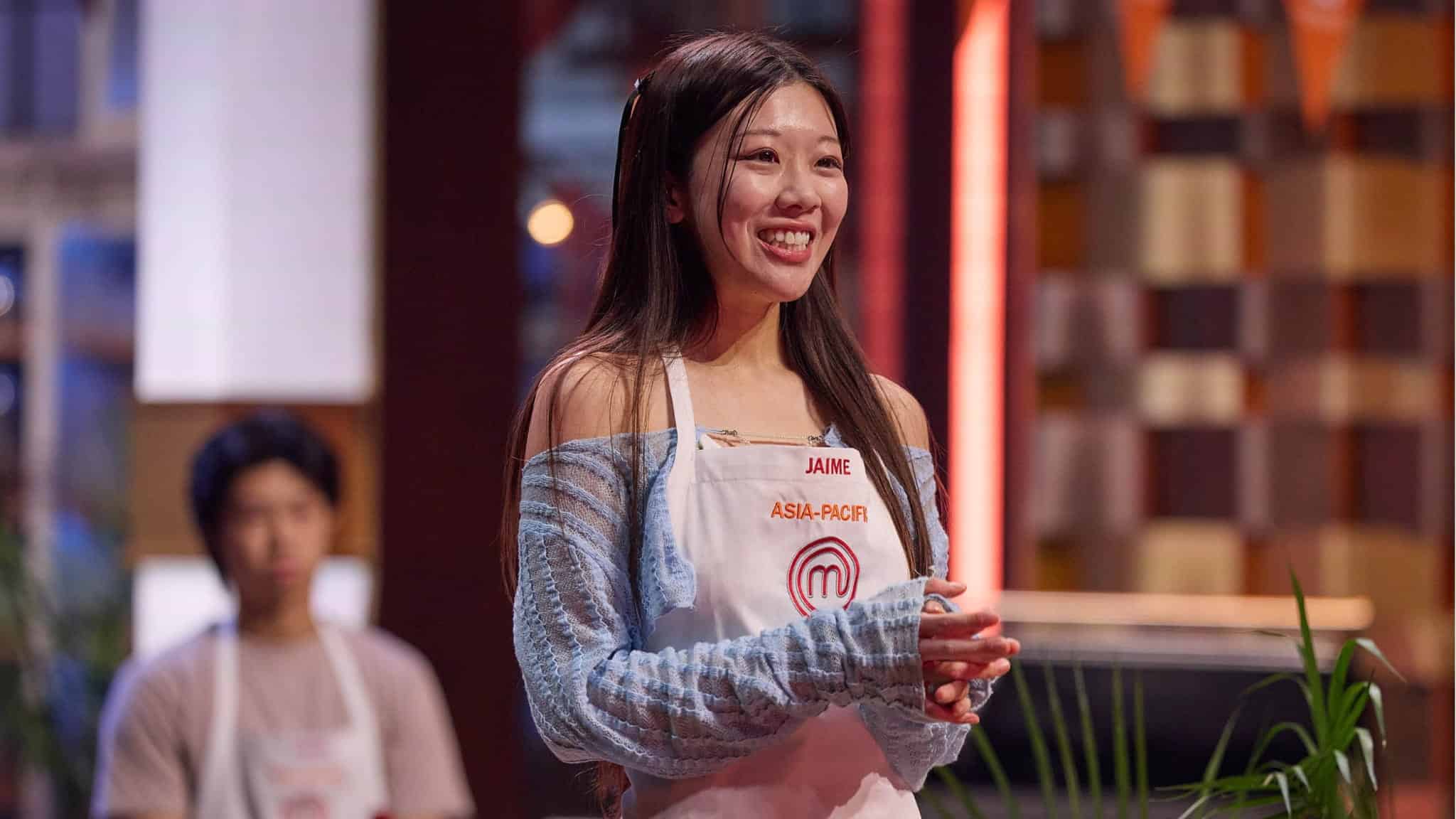 A smiling woman wearing a MasterChef Asia-Pacific apron stands indoors with hands clasped, radiating the confidence of a true Masterchef, while a blurred person sits in the background.