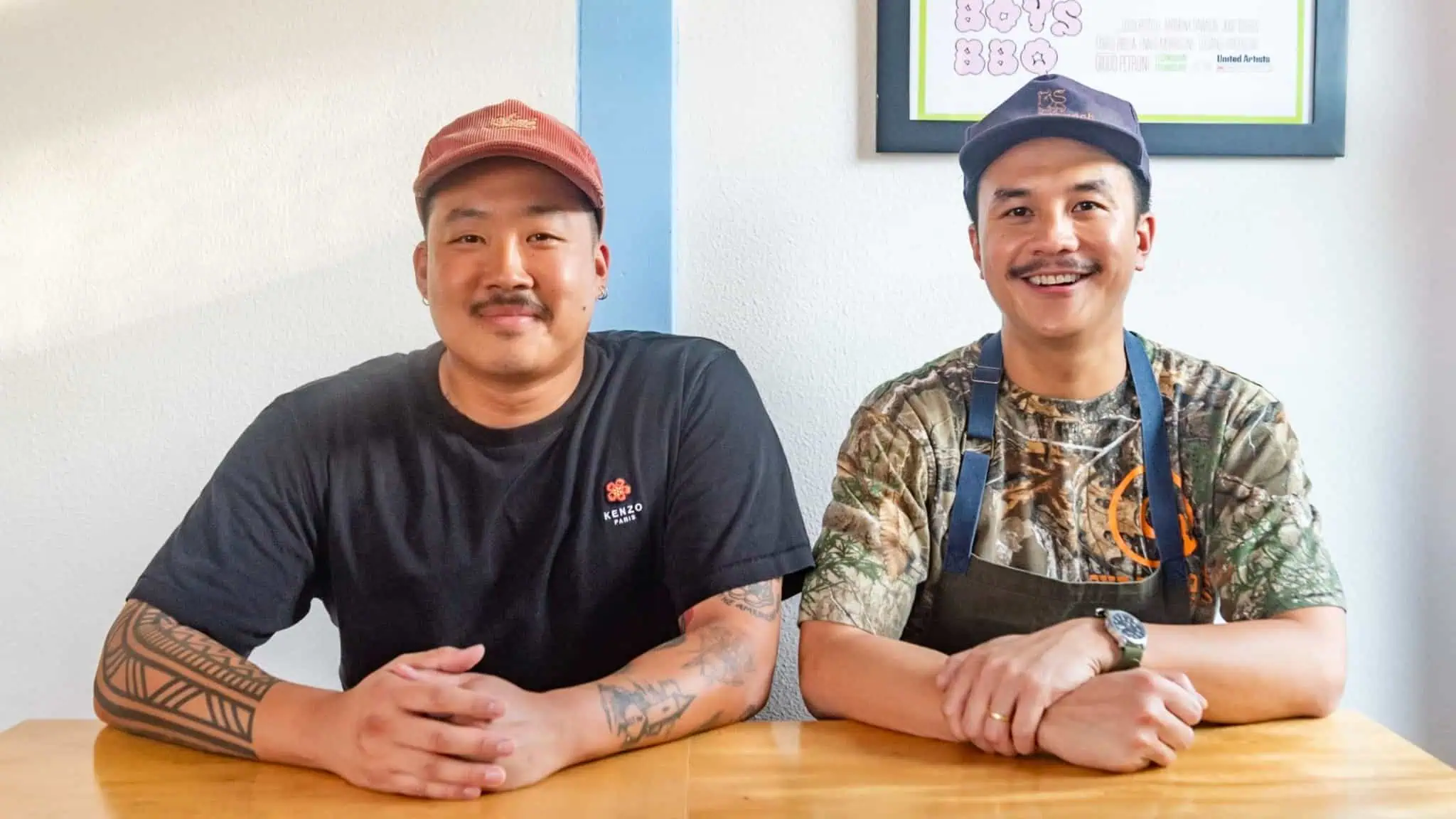 Two smiling men sit side by side at a wooden table. One wears a black t-shirt and red cap, the other has a camo shirt, apron, and a blue cap. Both have tattoos, and there’s a framed poster on the white wall behind them.
