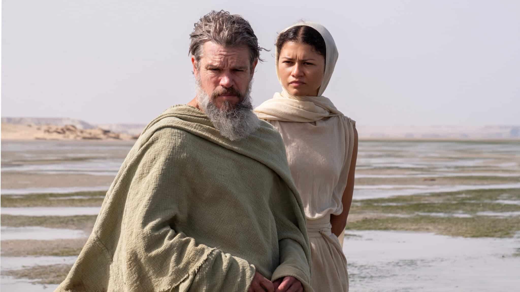 A bearded man in a green robe stands beside a woman in a cream headscarf and dress; both look serious and are outdoors by a vast, flat landscape with water and sand.