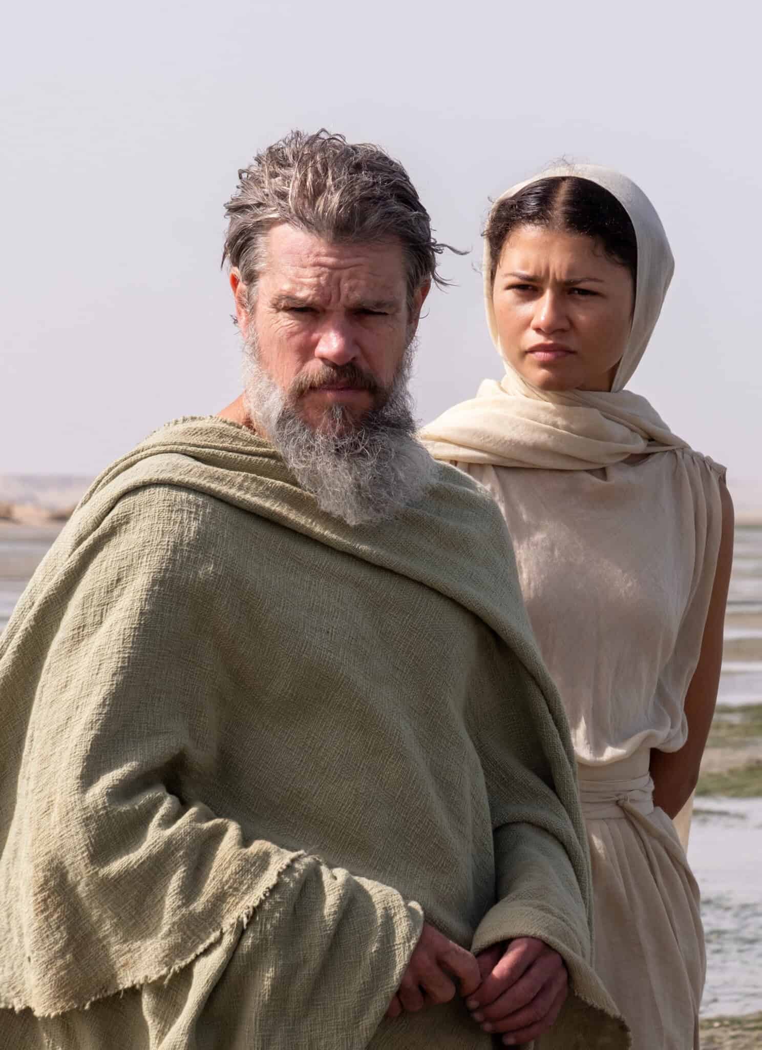 A bearded man in a green robe stands beside a woman in a cream headscarf and dress; both look serious and are outdoors by a vast, flat landscape with water and sand.