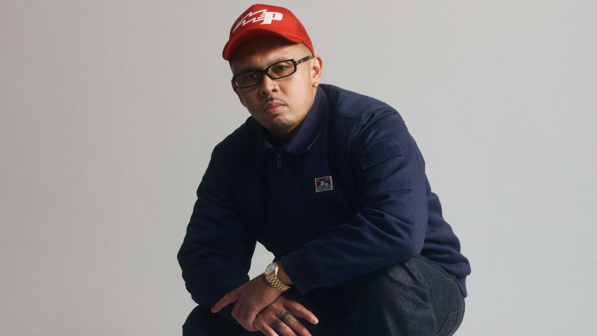 Man in glasses and a red cap squats in front of a plain light background, wearing a navy jacket, dark pants, a gold watch, and looking directly at the camera with a serious expression.
