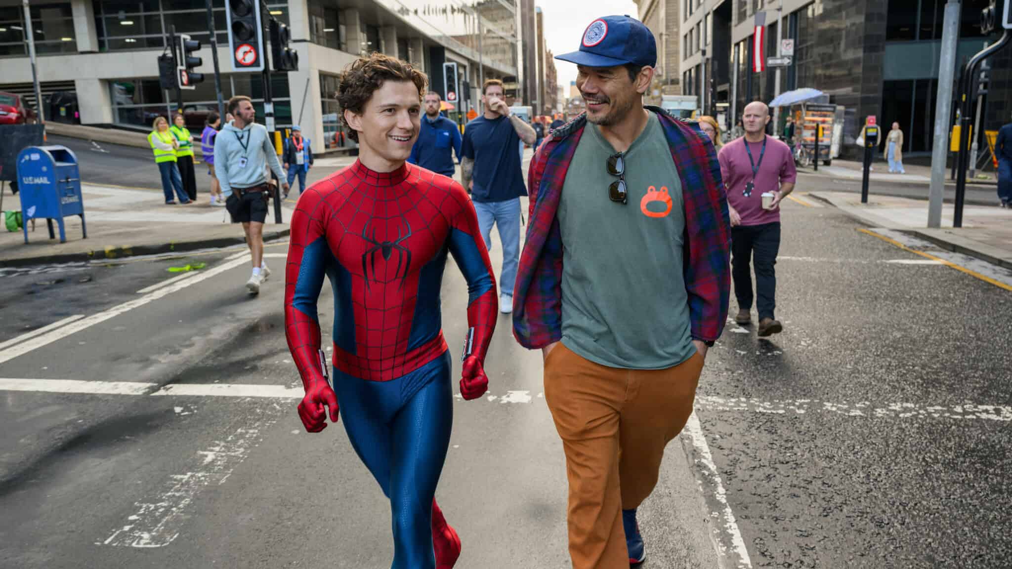 A person in a Spider-Man costume walks and smiles beside another casually dressed person on a city street, with people and buildings in the background, embodying the friendly neighborhood spirit of Spider-Man.