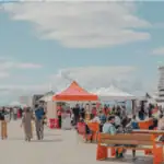People gather at an outdoor market with colorful tents on a sunny day. Some are seated on benches while others walk and browse stalls. A modern building is visible in the background under a partly cloudy sky.
