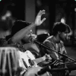 Two musicians perform on stage; one sings and plays guitar with a raised hand, while the other plays an instrument. Microphones and tabla drums are visible in the foreground. The image is in black and white.
