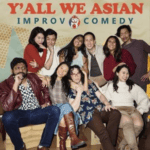 A group of ten people pose together and smile in front of a banner that reads “Y’ALL WE ASIAN IMPROV COMEDY.” They appear relaxed and cheerful, seated and standing in two rows.