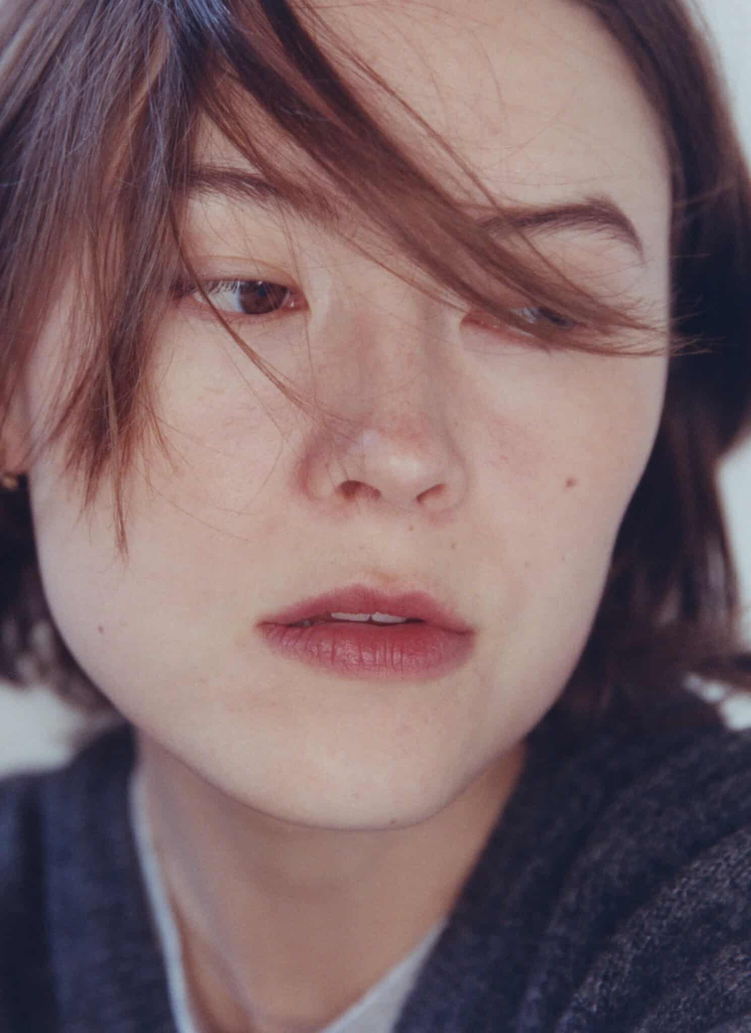 A person with short brown hair looks slightly away from the camera, lips parted. A lock of hair falls across their face, and the background is plain and light-colored.