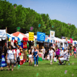 People walk and shop at an outdoor market with colorful vendor tents set up on a grassy area, surrounded by trees, on a sunny day. Some people are seated while others browse goods displayed at the stalls.