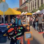 A colorful dragon costume is worn at a lively outdoor market with yellow tents, food stands, and people walking and socializing under sunny skies.