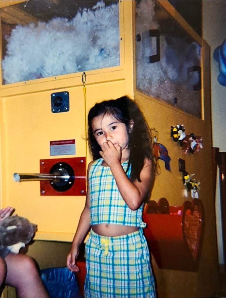 A young Wasian girl in a plaid outfit stands indoors, sucking her thumb near a large yellow machine filled with white stuffing and decorated with stickers.