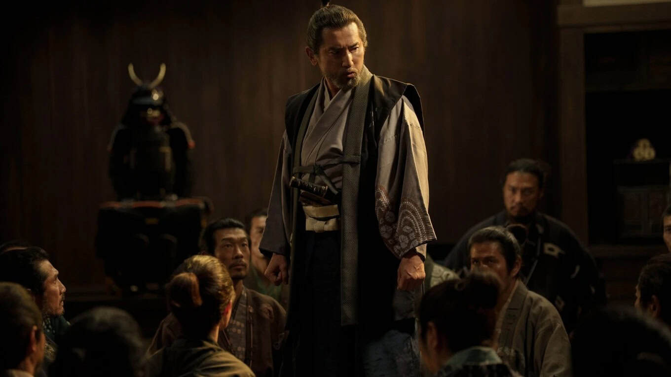 A man in traditional Japanese clothing stands and addresses a seated group in a dimly lit room, with a samurai armor display visible in the background.