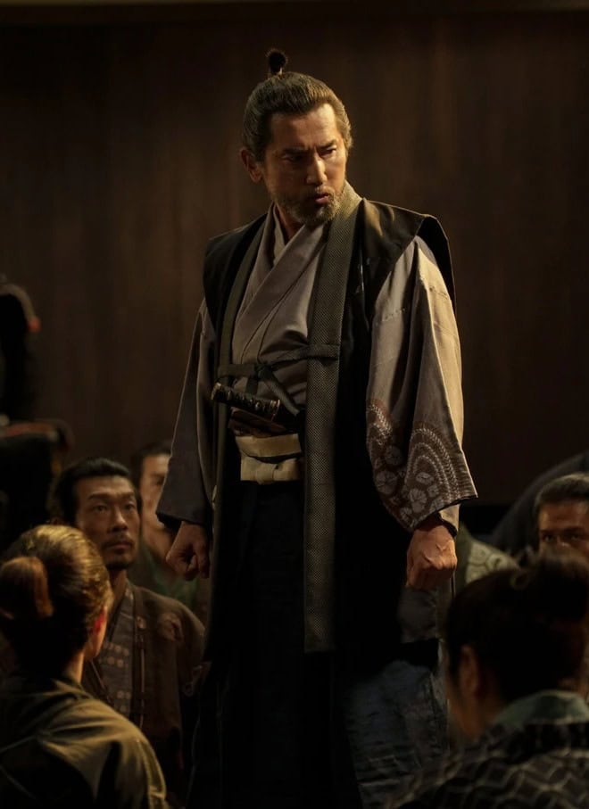 A man in traditional Japanese clothing stands and addresses a seated group in a dimly lit room, with a samurai armor display visible in the background.