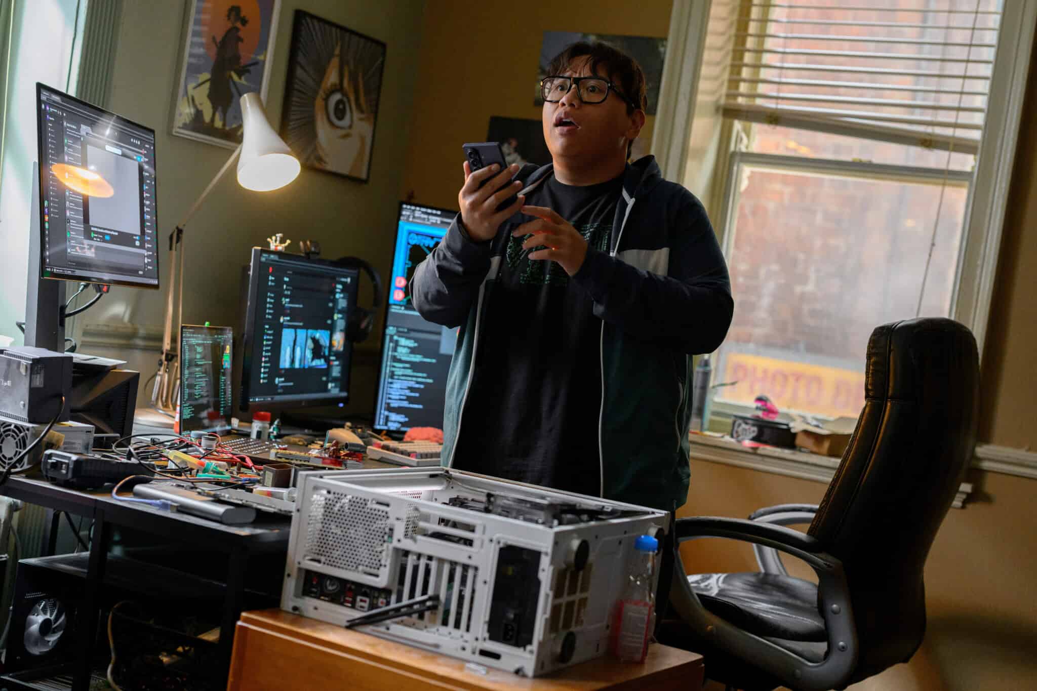 A person stands in a cluttered room with computer parts, holding a phone and looking surprised. Multiple monitors display code, and sunlight streams through a window. The scene suggests tech work or troubleshooting.