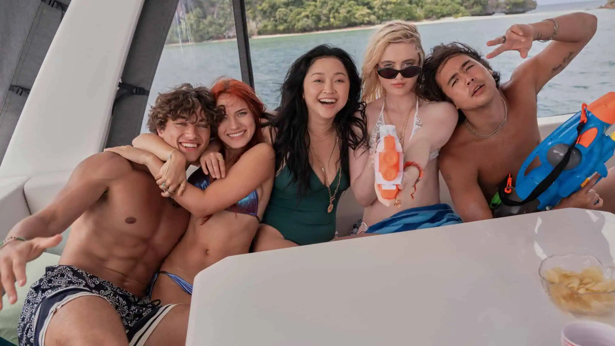 Five young adults in swimwear smile and pose on a boat. Two hold water guns, and a tropical landscape is visible in the background. Everyone appears to be having fun.