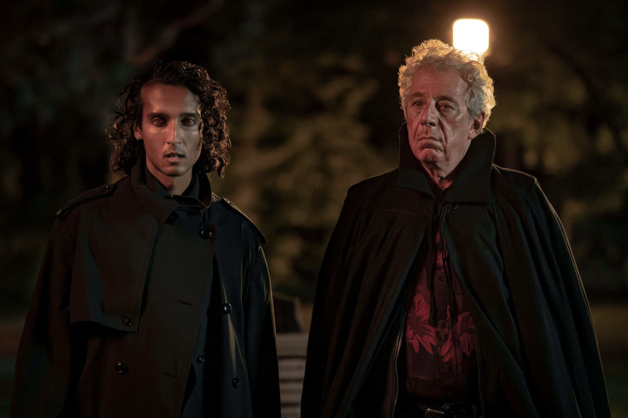 Two men in dark coats stand outside at night under a streetlamp; one has curly hair and a serious expression, the other has gray hair and appears solemn. Trees and dim lighting fill the background.
