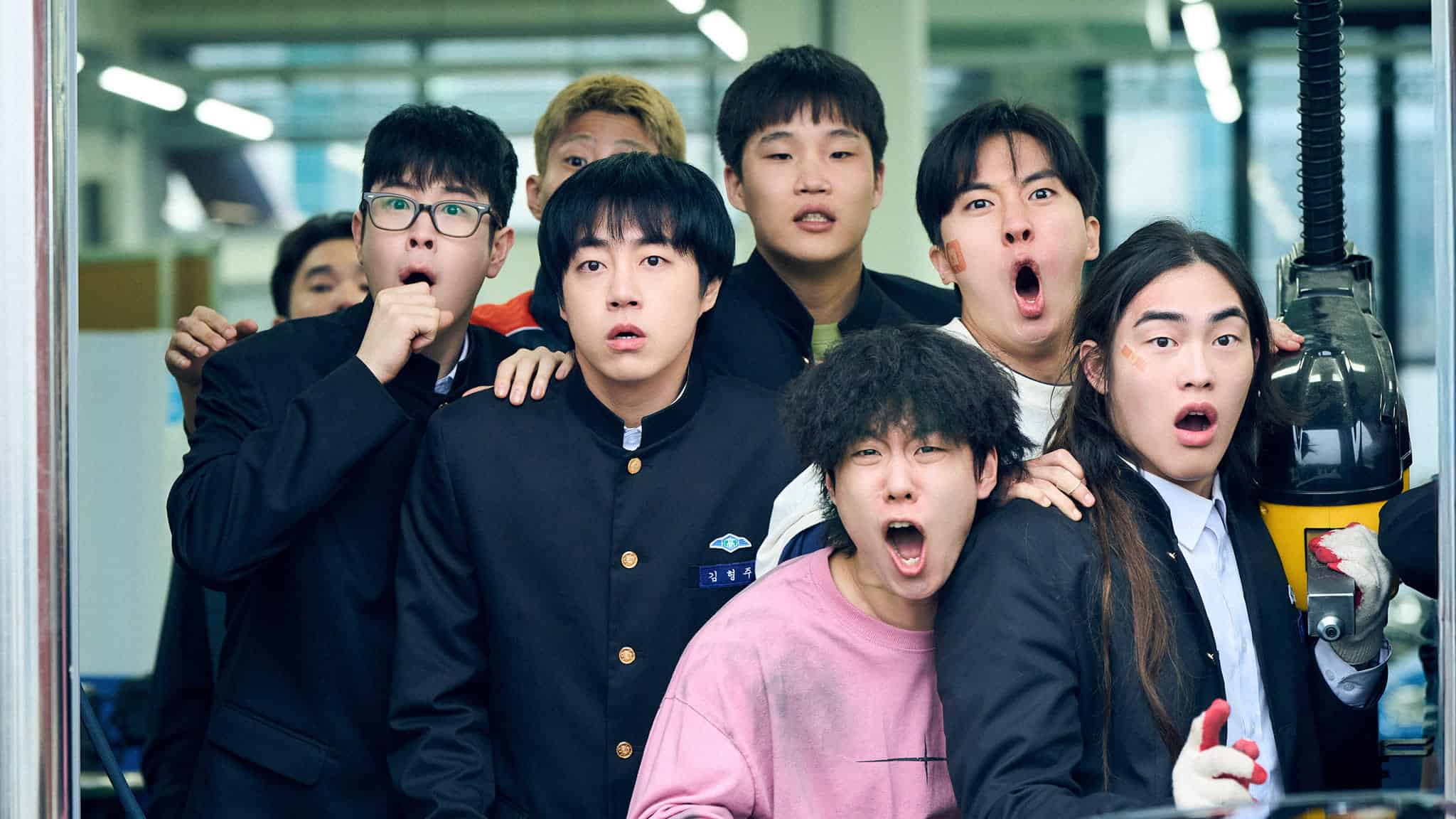 A group of seven shocked young people, most wearing school uniforms, stand close together indoors, staring intensely at something off-camera.