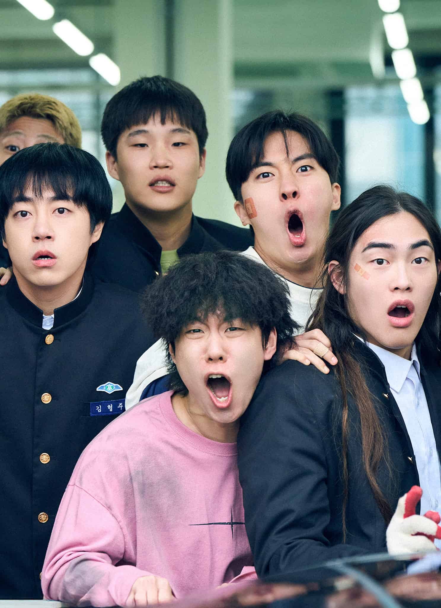 A group of seven shocked young people, most wearing school uniforms, stand close together indoors, staring intensely at something off-camera.