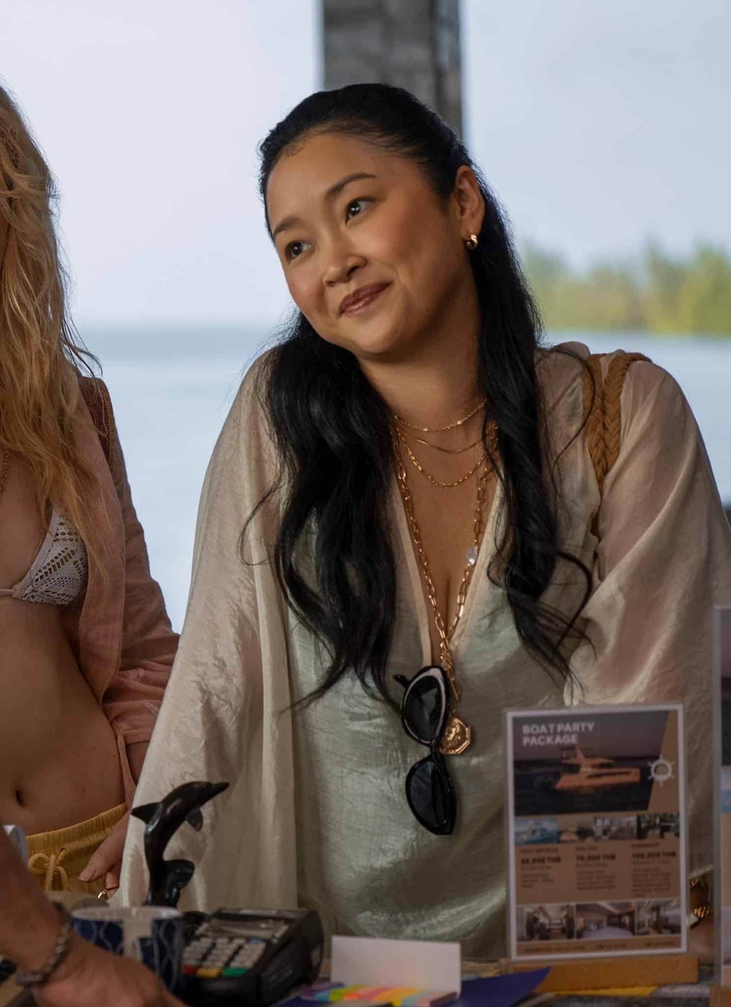 Two women stand at a counter, one in a bikini top and open shirt, the other in a light blouse, smiling at a man in glasses and a cap, with a beach scene visible in the background.