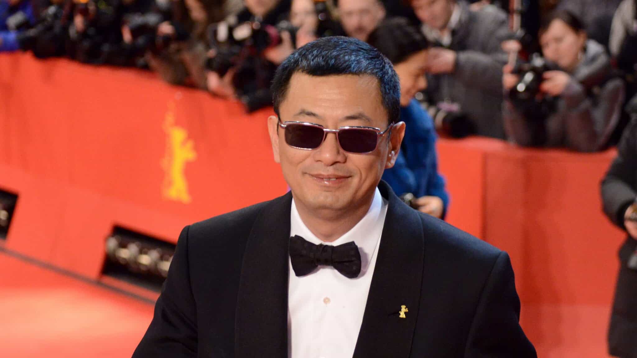A man in a tuxedo and sunglasses smiles on a red carpet with photographers in the background.
